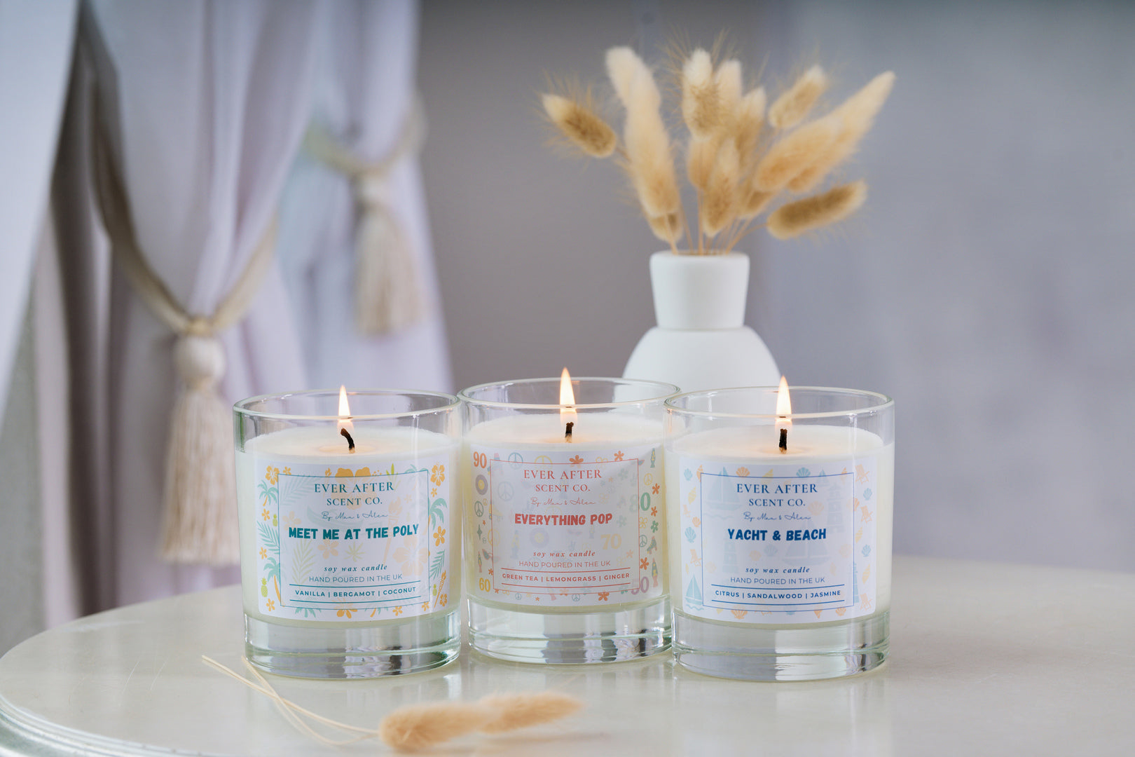 Ever After Scent Co Three lit candles with labels on a table, accompanied by a vase with dried plants.