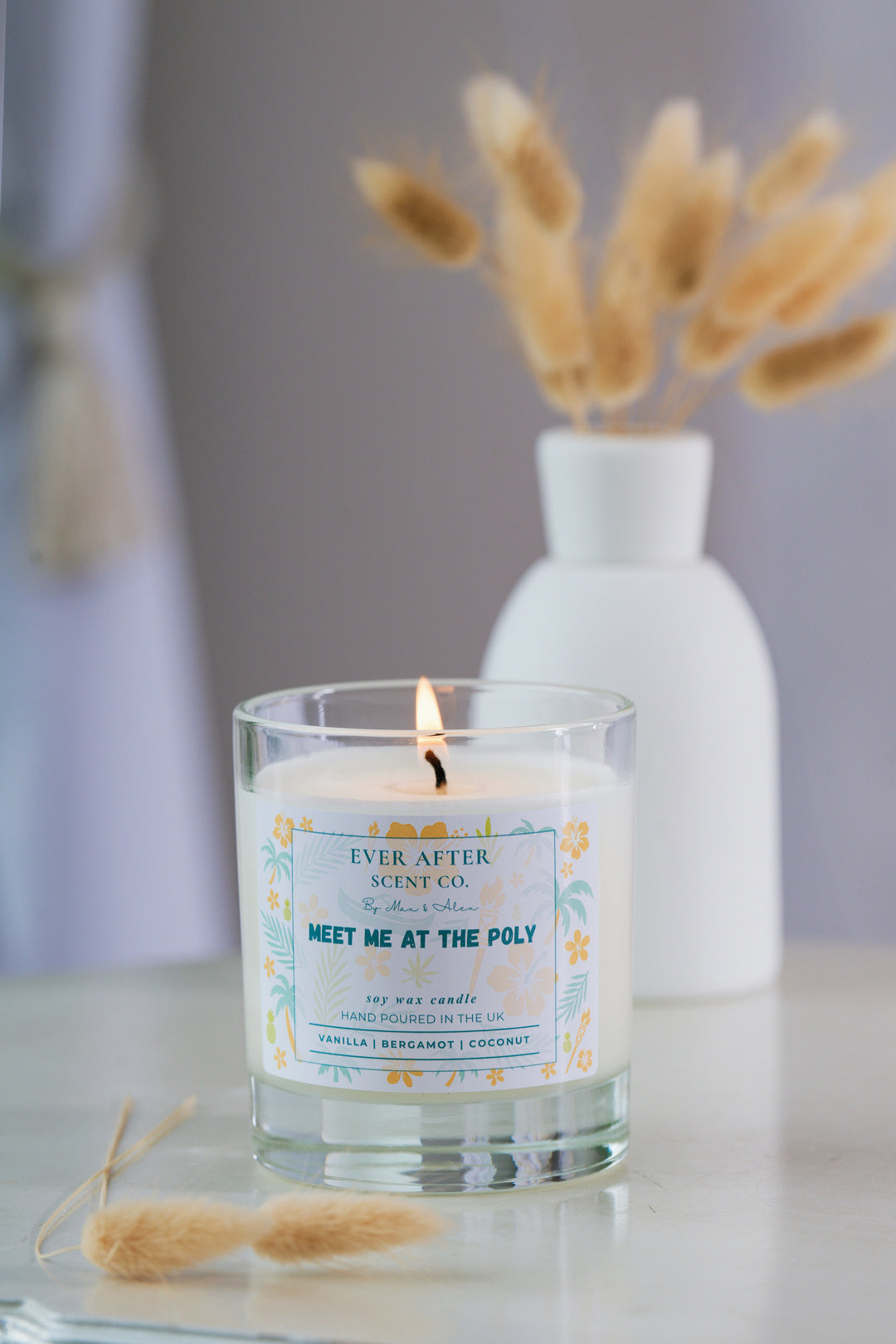 Ever after scent co meet me at the poly Candle with a label in a glass holder on a surface with a blurred background