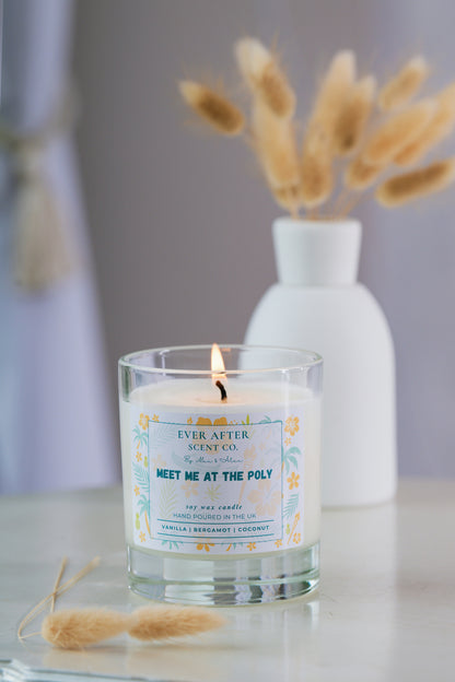 Ever after scent co meet me at the poly Candle with a label in a glass holder on a surface with a blurred background