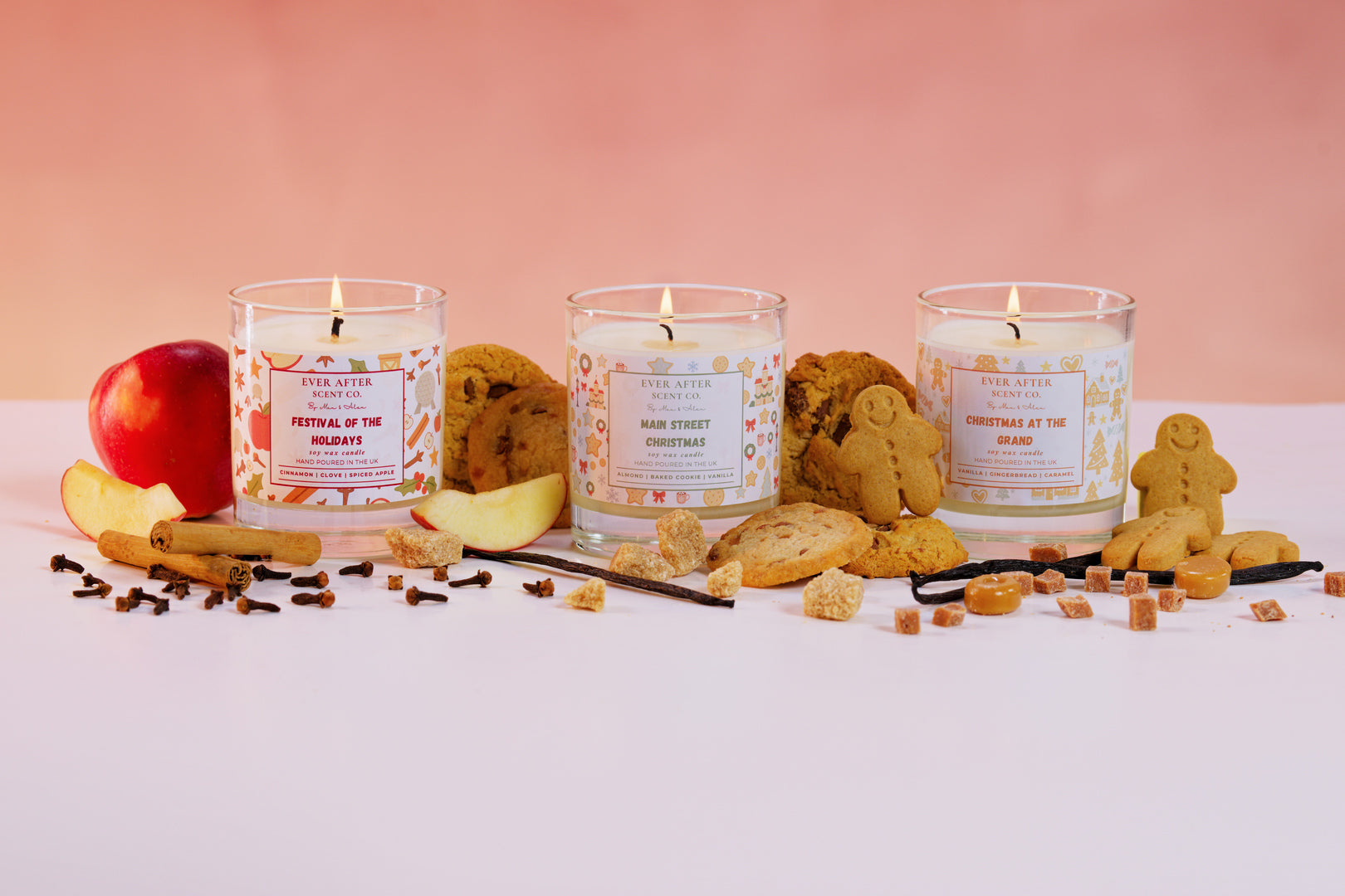 Three candles with festive labels on a white surface with cookies and apples.